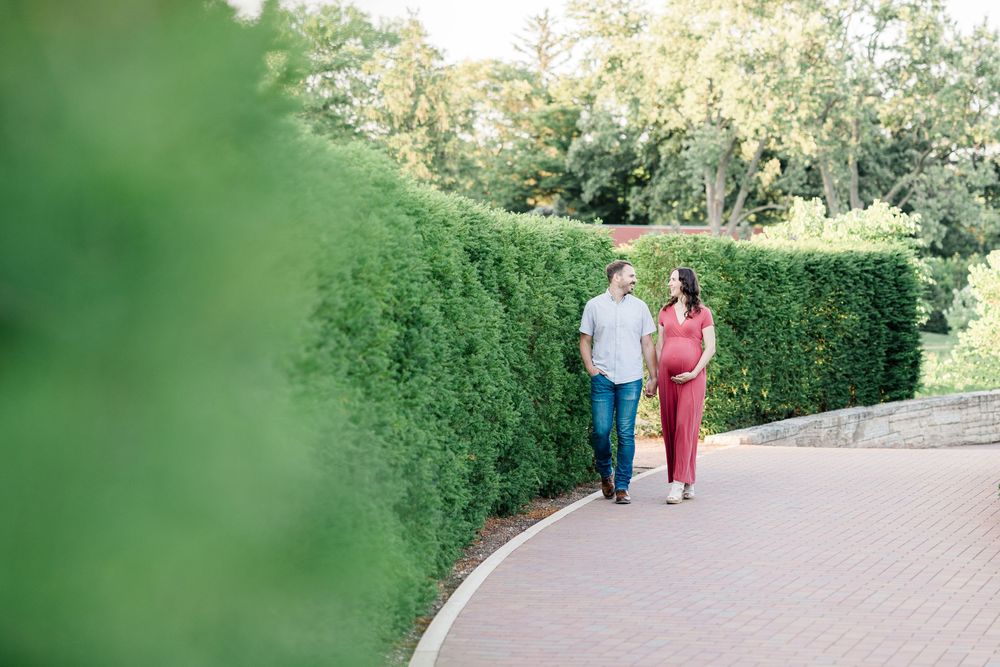 Expectant couple strolling along a brick path beside tall green hedges in a sunny garden.