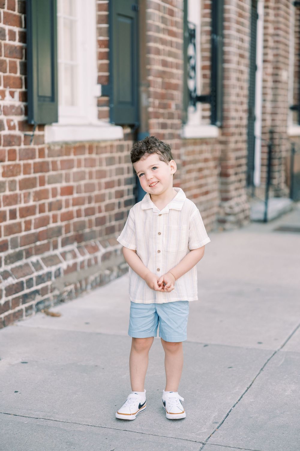 Adorable young boy in cream shirt and blue shorts smiling in front of brick building.