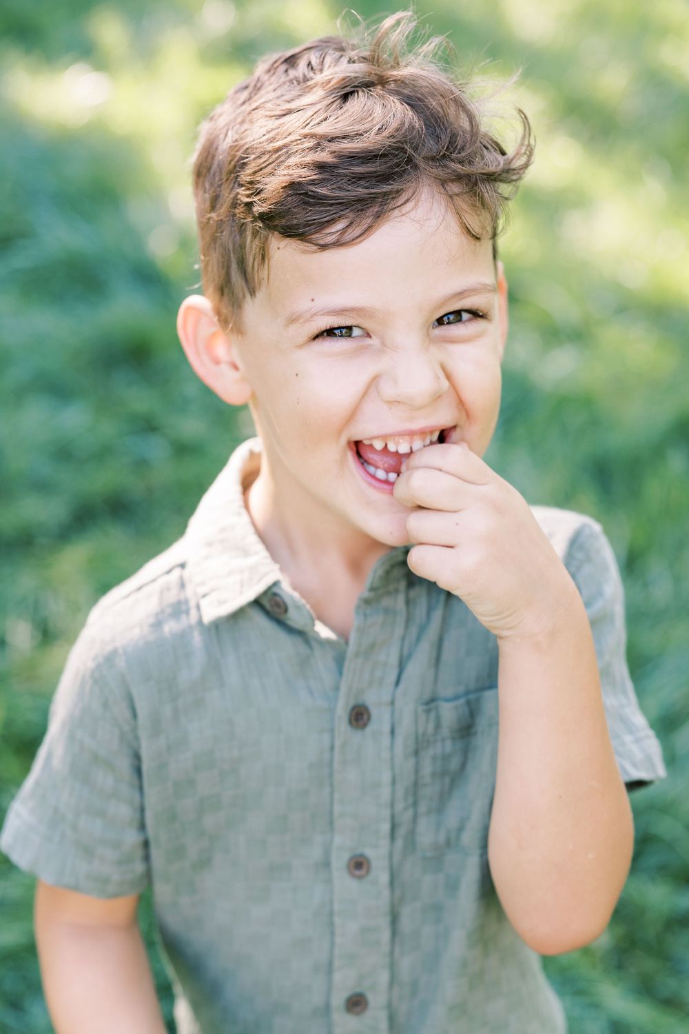 Happy young boy with curly hair laughing outdoors in green field wearing gray shirt.
