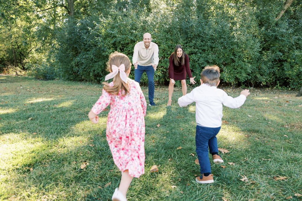 Family playing together on green lawn, two children running toward parents in wooded setting.