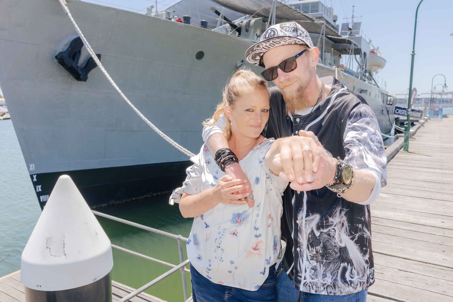 Couple embracing on a sunny harbor dock near a naval ship, man pointing at camera wearing bandana and graphic tee.
