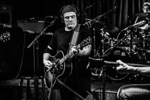 Black and white photo of a male guitarist performing on stage with an acoustic guitar and microphone.