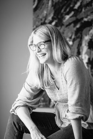 Black and white portrait of a smiling blonde woman wearing glasses, sitting casually in a relaxed, candid pose.