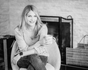 Black and white photo of a smiling blonde woman sitting in a chair holding a mug near a fireplace.