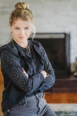 Blonde woman in navy eyelet blouse and jeans poses with arms crossed by a fireplace.
