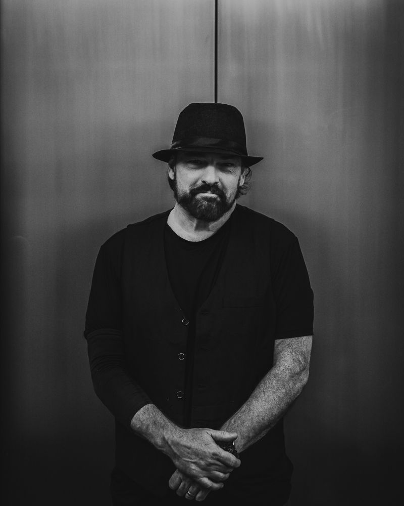 Black and white portrait of a bearded man in a black hat and outfit, arms crossed, standing against a metallic wall.