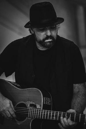 Black and white photo of a bearded man in a fedora hat playing an acoustic guitar, looking down thoughtfully.
