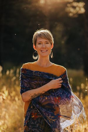 Smiling woman with short hair wearing mustard top and floral blue scarf, standing in golden sunlit field.