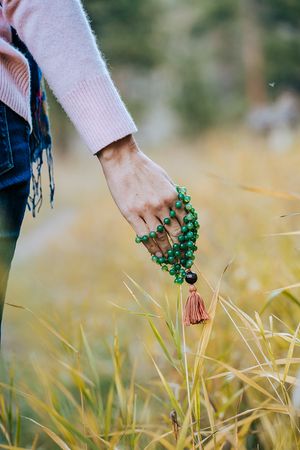 Hand holding green mala beads with tassel over golden grass field in nature.