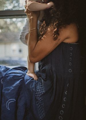 Two people embracing tenderly near a window, one wearing a navy blue button-back dress with eyelet details.