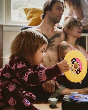 A young child in a purple patterned outfit holds a yellow vinyl record while adults with tattoos relax in the background.