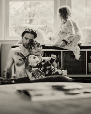 Black and white photo of a shirtless tattooed man and child relaxing indoors, with another child playing in the background.