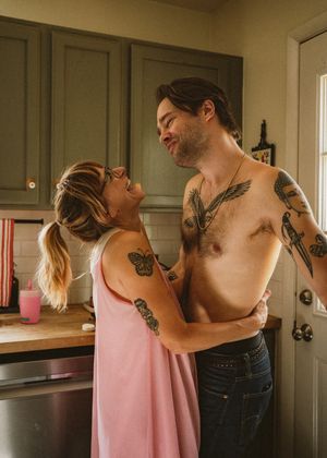 Tattooed couple embracing and laughing together in a cozy kitchen, woman in pink dress, man shirtless in jeans.