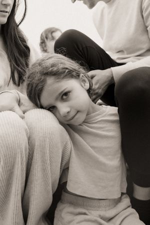 Black and white photo of a young girl resting her head on an adult's knee, smiling softly while seated among family members.