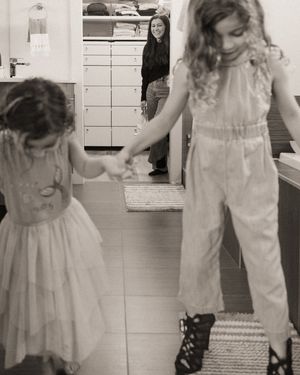 Two young girls hold hands and dance in a room while a smiling woman watches from the doorway in this black and white photo.