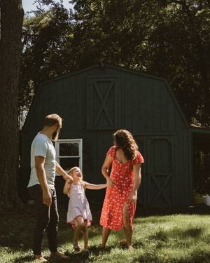 A family of three holds hands in a sunny backyard, with a dark barn and lush trees in the background.