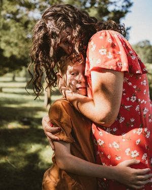 A mother with curly hair in a red floral dress hugs a young child outdoors in a sunny green park.