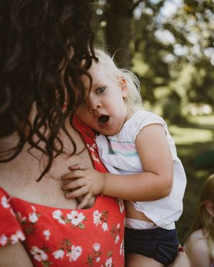 A surprised toddler with blonde hair being held by a curly-haired woman in a red floral dress outdoors.