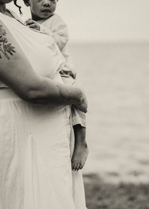 Black and white photo of a pregnant woman in white dress holding a child by the ocean, showcasing a tattoo on her arm.