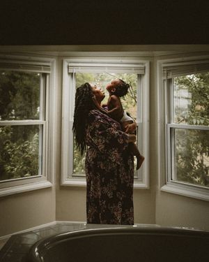 A mother lifts her baby joyfully near a bay window with lush greenery, wearing a floral dress with long braids.