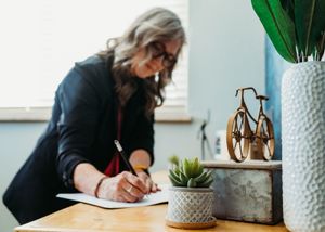Woman with gray hair writing in notebook at desk with succulent plant and bicycle decor in bright office.