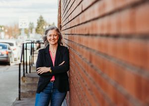Smiling woman with gray hair leaning against a brick wall outdoors, wearing a black blazer and jeans.