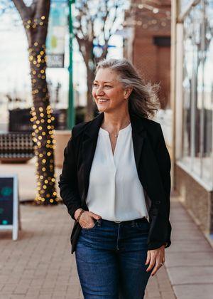 Smiling middle-aged woman with gray hair wearing a black blazer and white blouse walking outdoors.