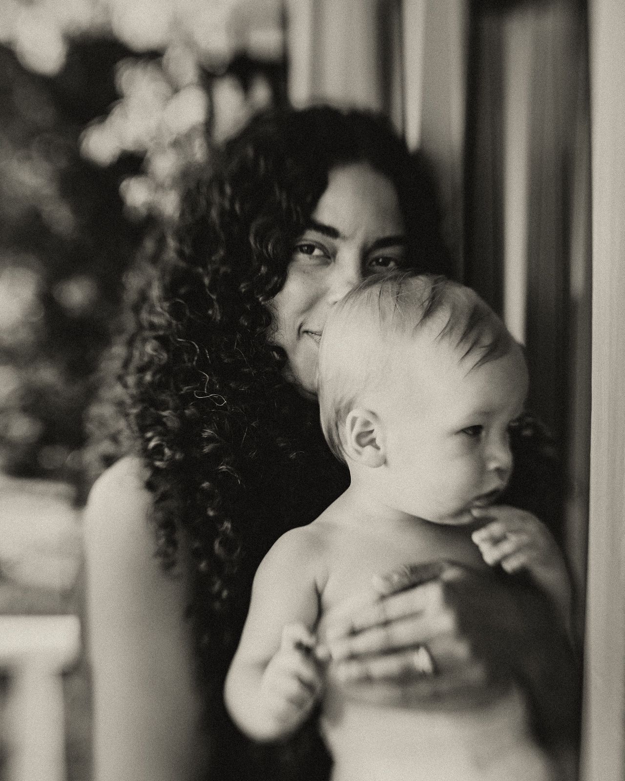 Black and white photo of a curly-haired woman tenderly holding a baby near a window.