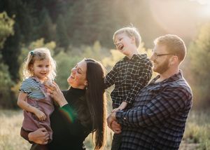Happy family with two young children laughing outdoors in a sunlit meadow, parents holding kids in warm golden light.
