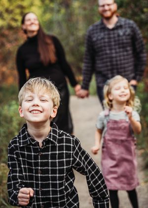 A joyful family of four walks on a path outdoors, with a smiling boy in a plaid shirt leading the way.