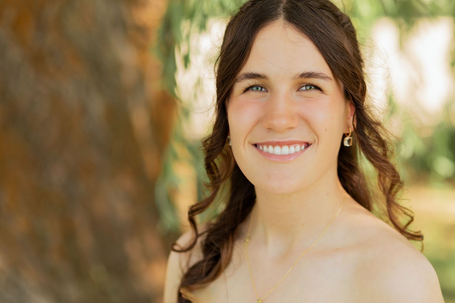 Smiling young woman with curly brown hair posing outdoors near a tree in warm sunlight.