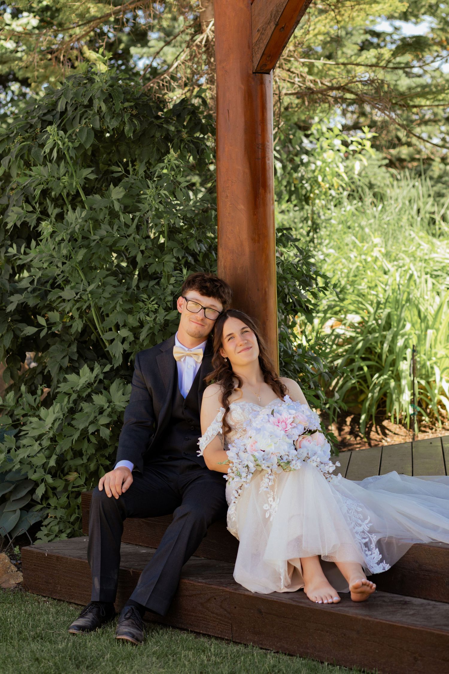 Bride and groom sit together on wooden steps under a pergola surrounded by lush greenery on their wedding day.