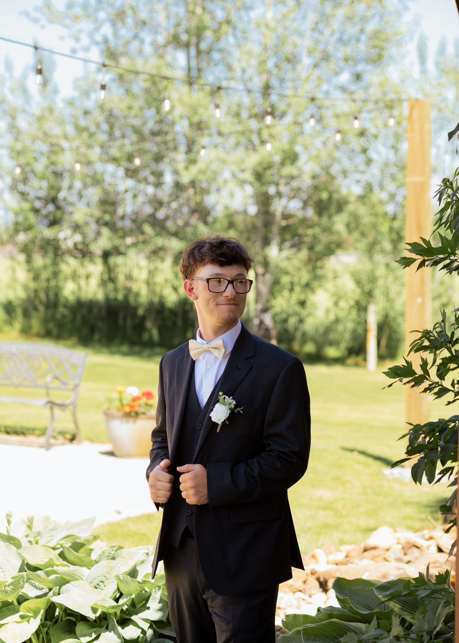 Young groomsman in a dark suit with bow tie and boutonniere stands outdoors at a sunny wedding venue.