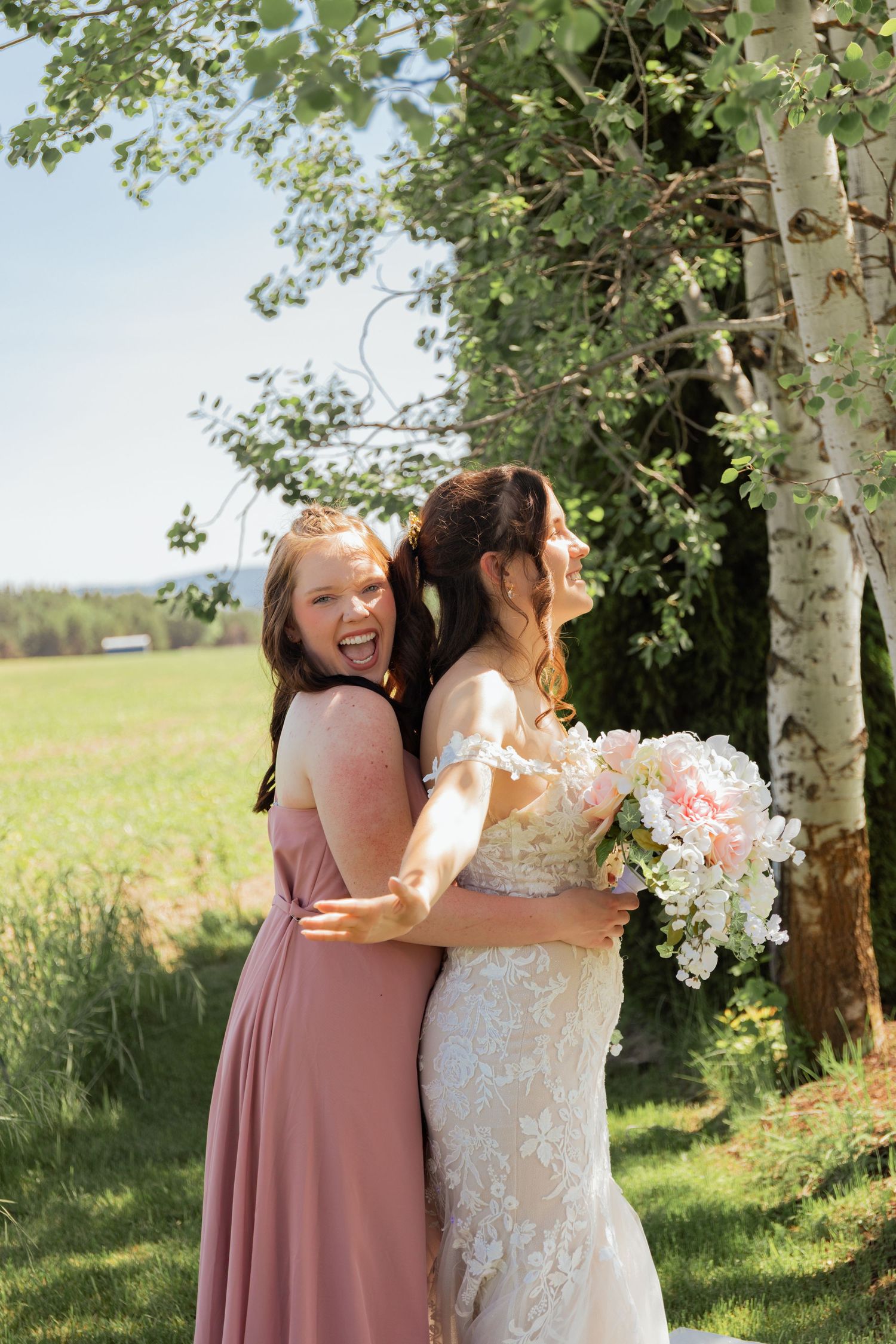 Bridesmaid in dusty rose gown hugs smiling bride holding pink and white bouquet near birch trees outdoors.