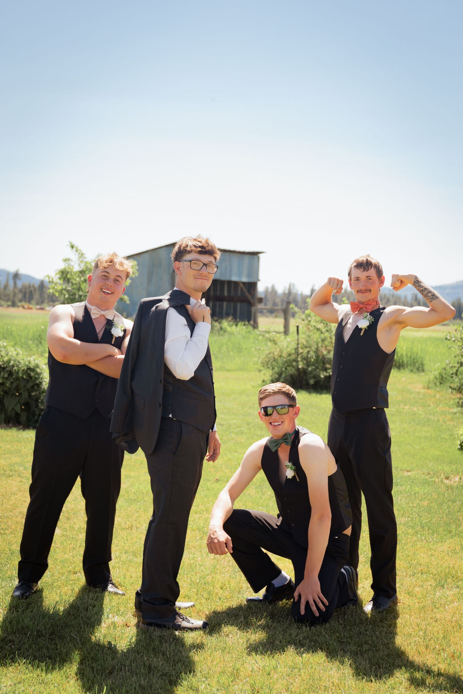 Four groomsmen in black vests pose playfully on green grass with a rustic barn backdrop on a sunny wedding day.