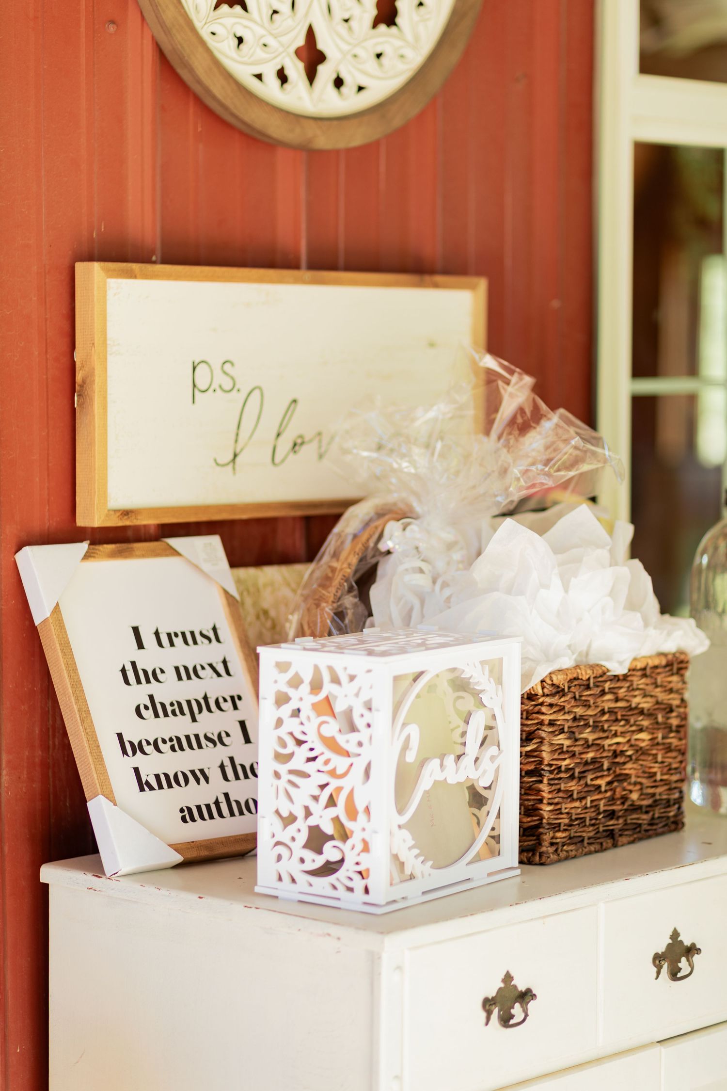 Wedding gift table with white card box, wicker basket, and inspirational signs on white dresser against red wall.
