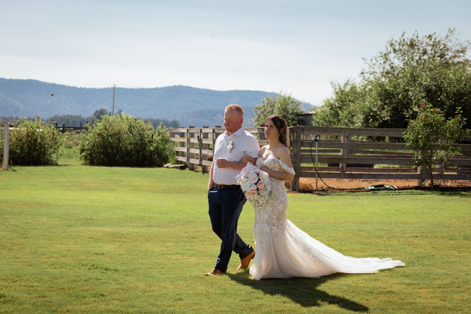 Bride in lace gown and groom in white shirt walk hand in hand on green lawn with mountain backdrop.