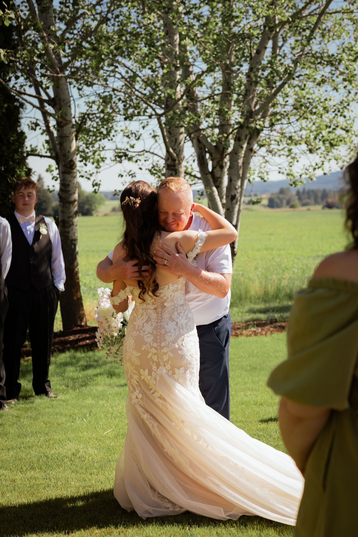 Bride in lace mermaid gown shares emotional hug outdoors on her wedding day surrounded by guests.