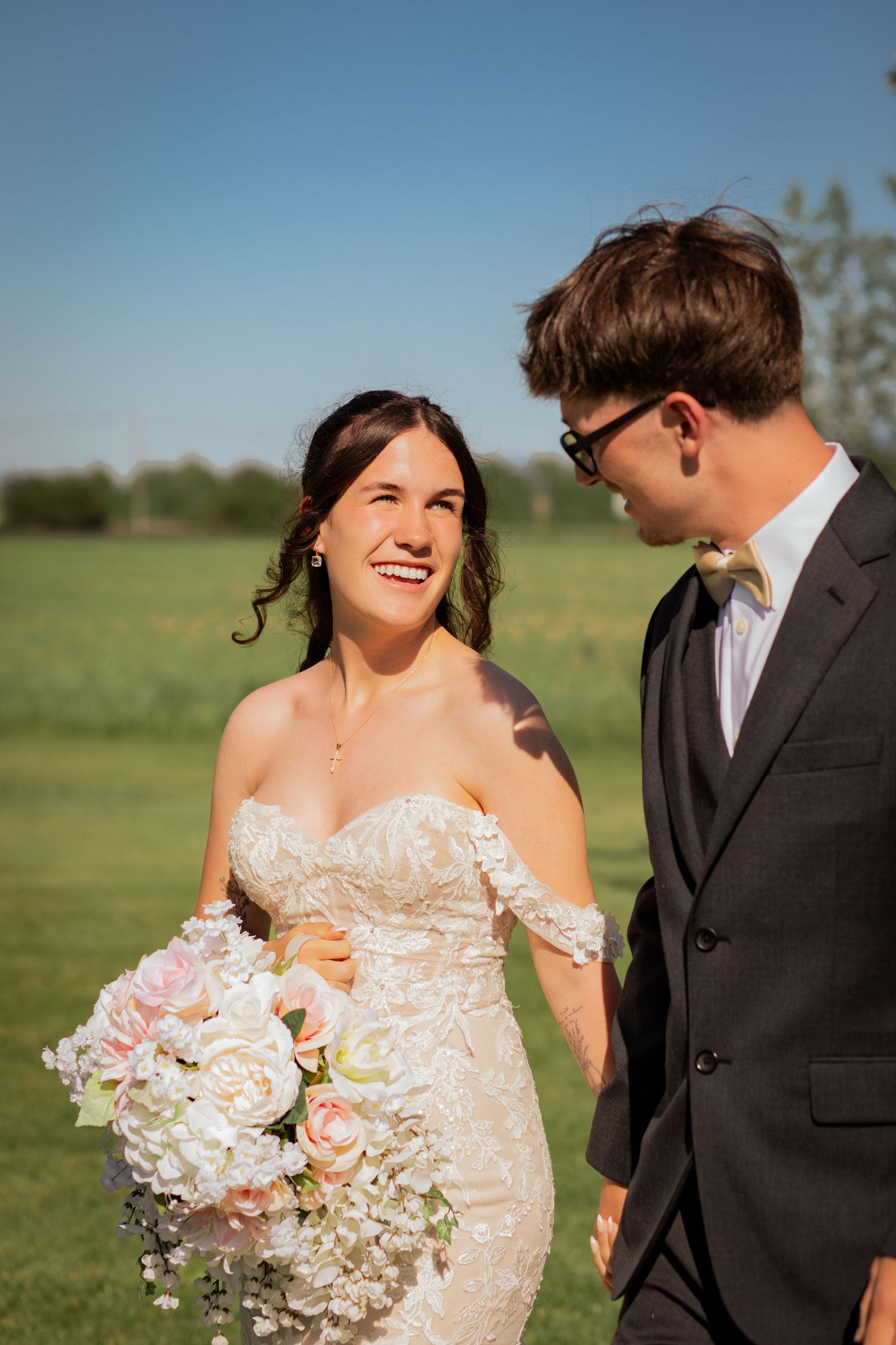 Smiling bride in lace off-shoulder gown holds pastel bouquet while walking with groom in dark suit outdoors.