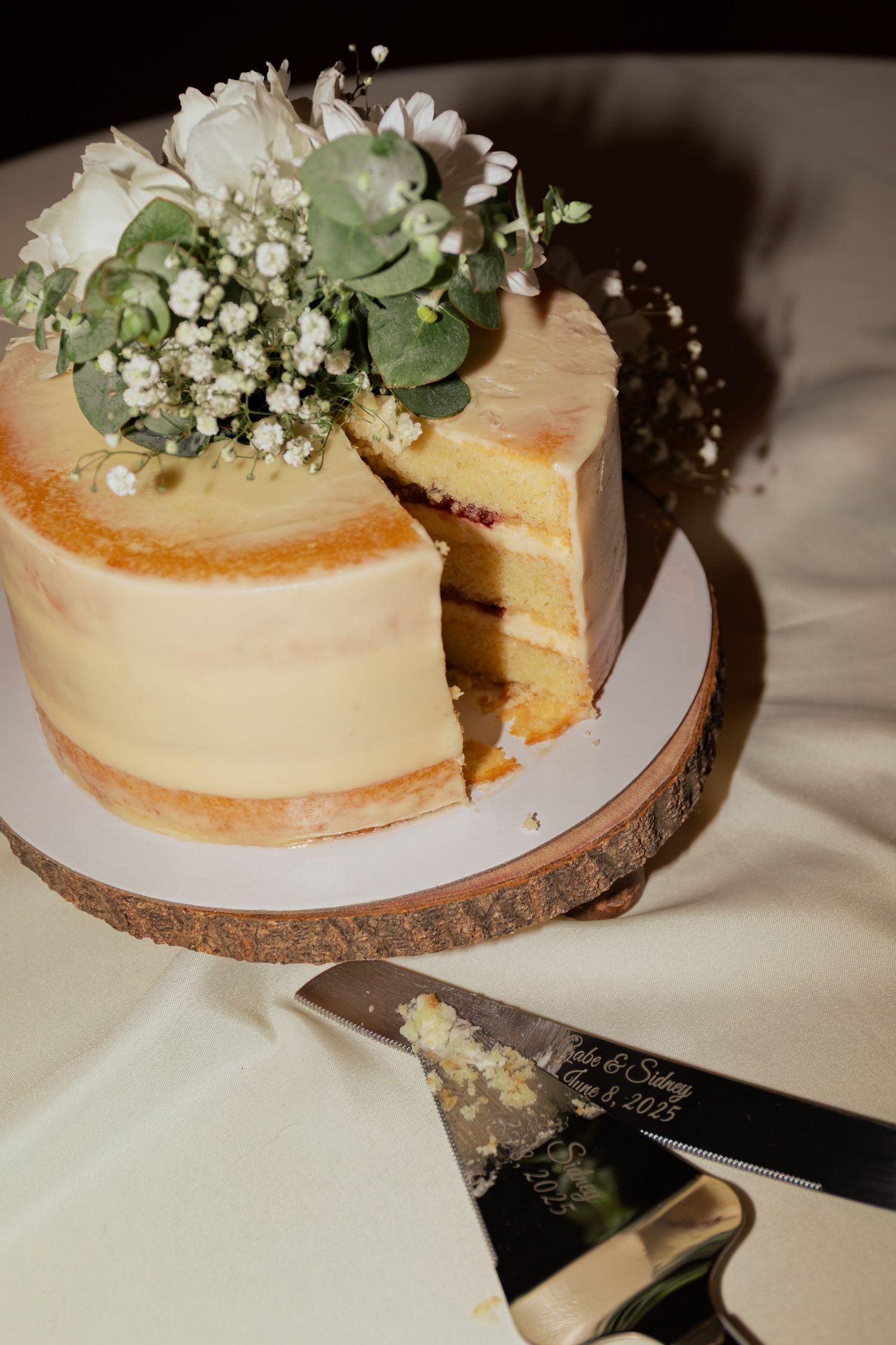 Single-tier naked wedding cake topped with white flowers and greenery, displayed on a rustic wood slice cake stand.