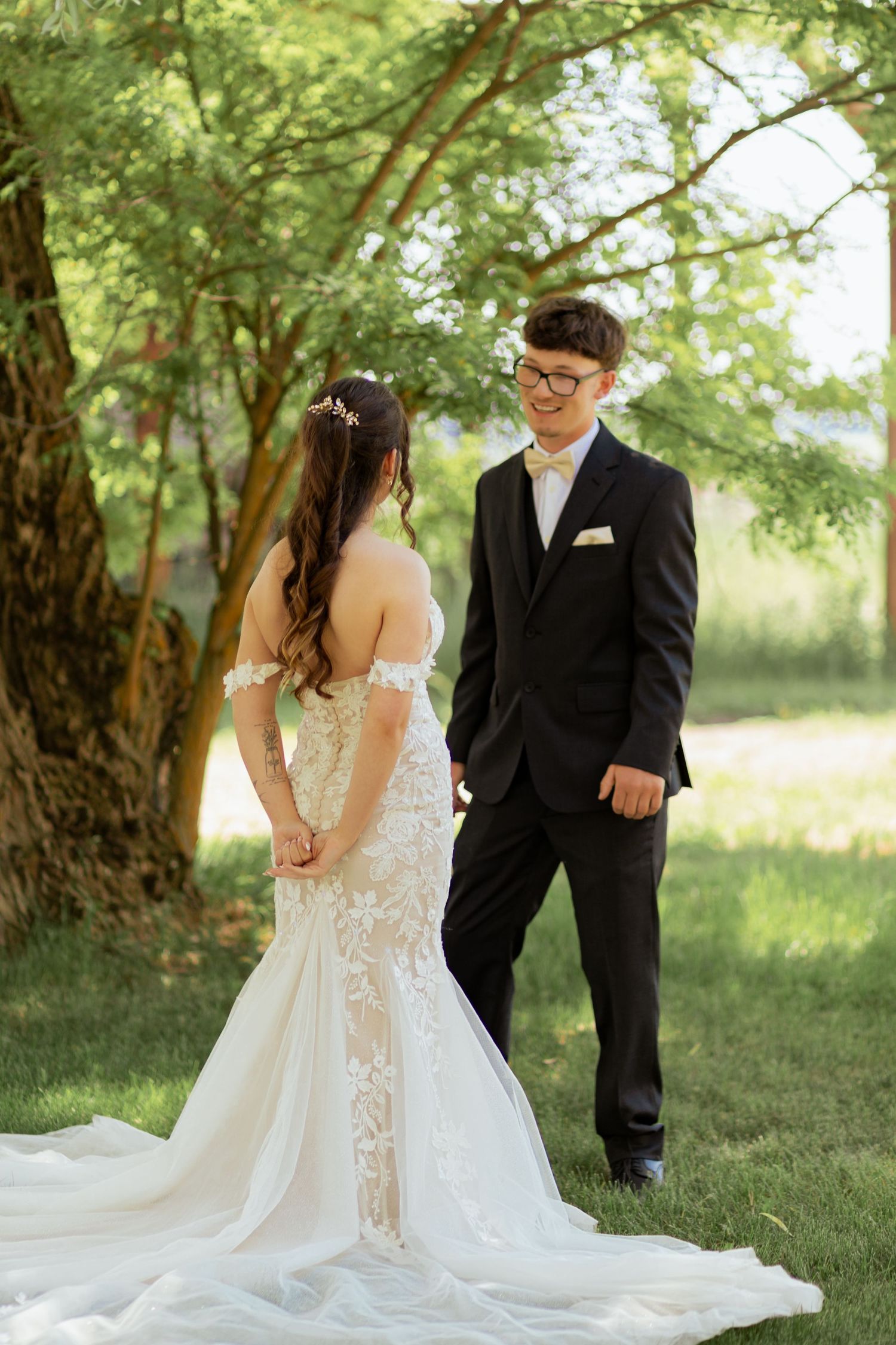 Bride in lace mermaid gown and groom in black suit share a romantic moment outdoors under green trees.