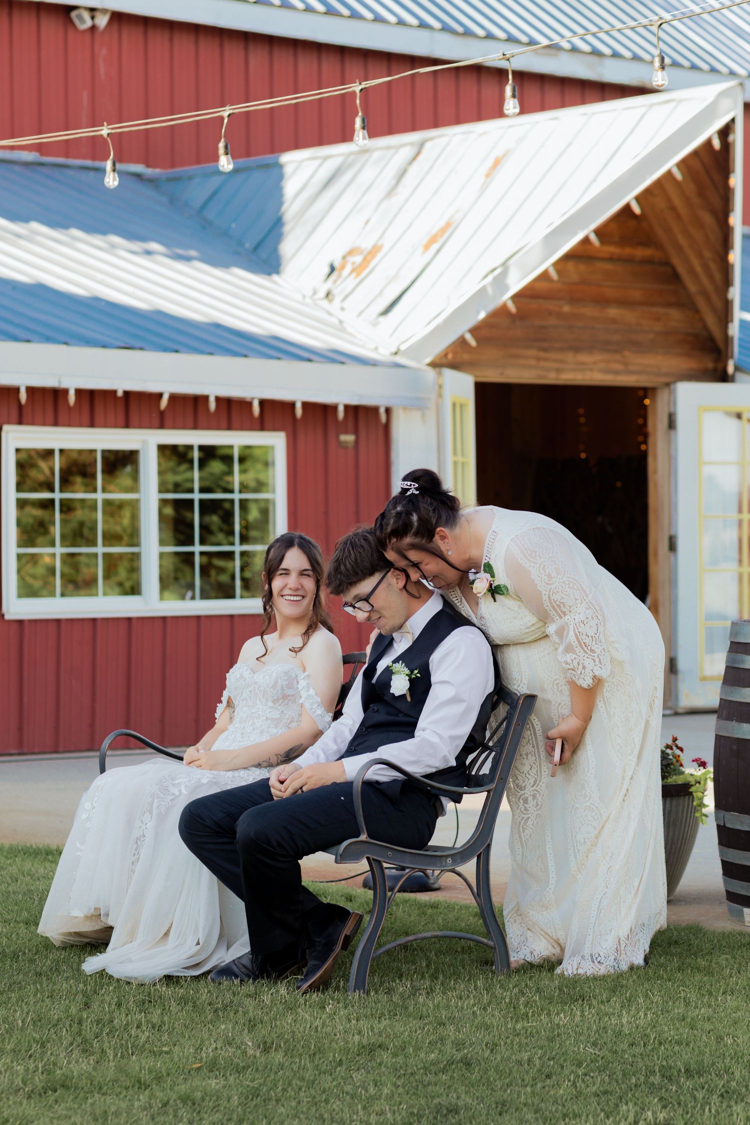 Two brides share a tender moment on a bench outside a red barn wedding venue at their outdoor reception.