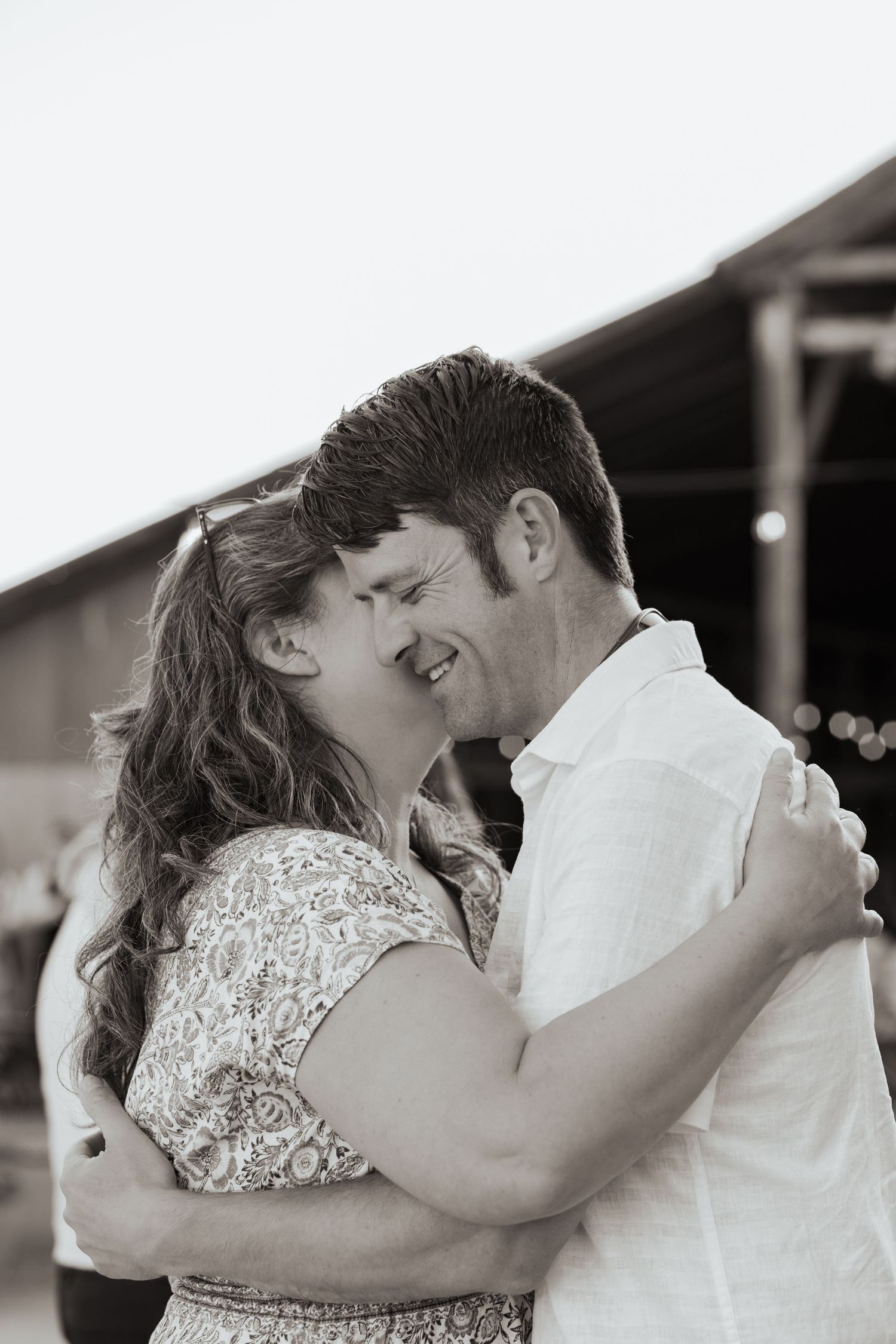 Black and white photo of a couple slow dancing and smiling at each other outdoors near a rustic barn.
