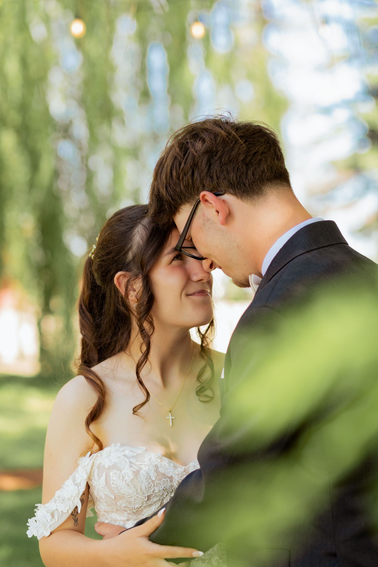 A bride and groom share an intimate moment outdoors, nearly kissing under a willow tree with soft bokeh lighting.