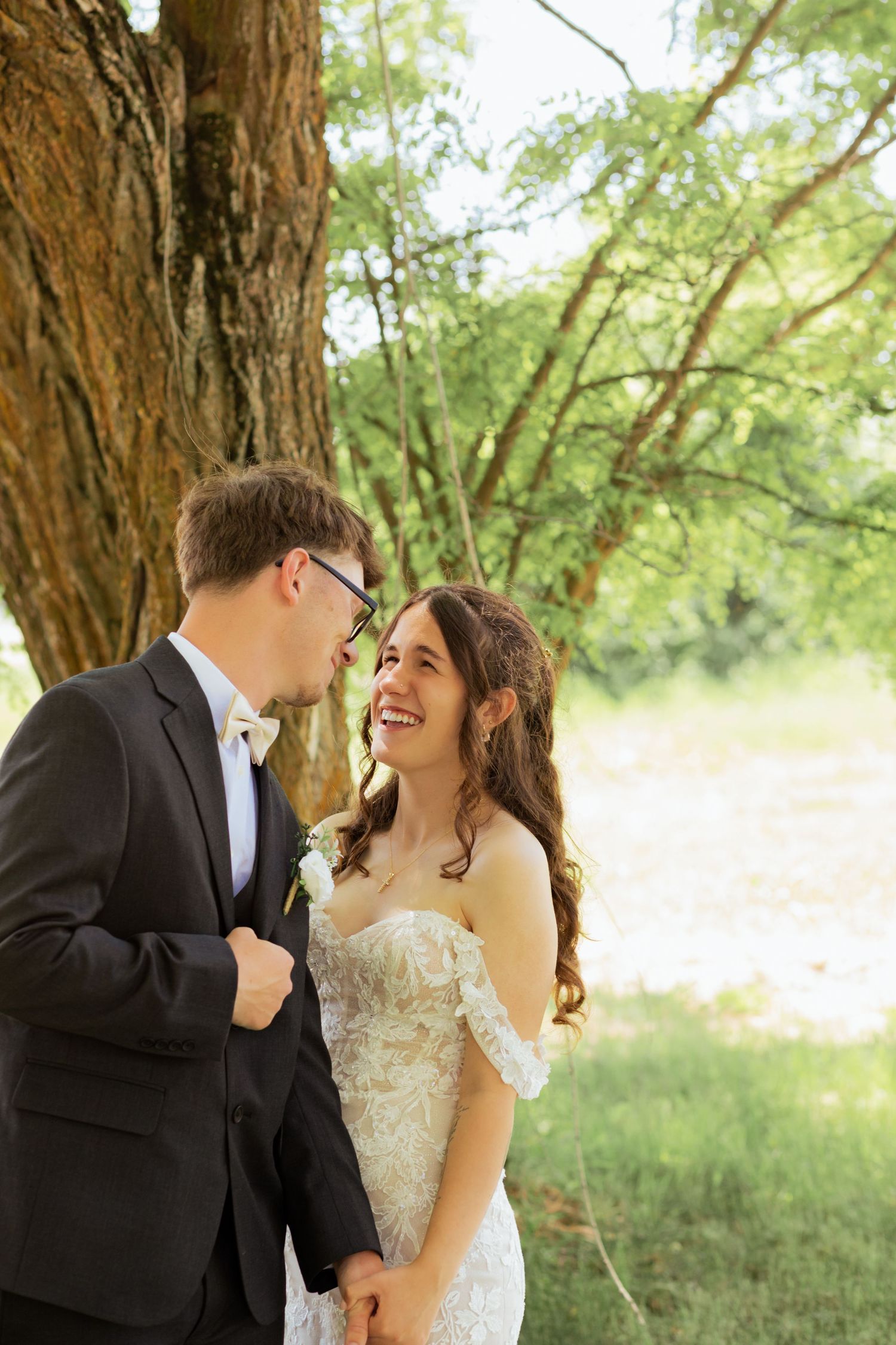 Bride and groom share a joyful moment laughing together under a large tree in an outdoor wedding setting.