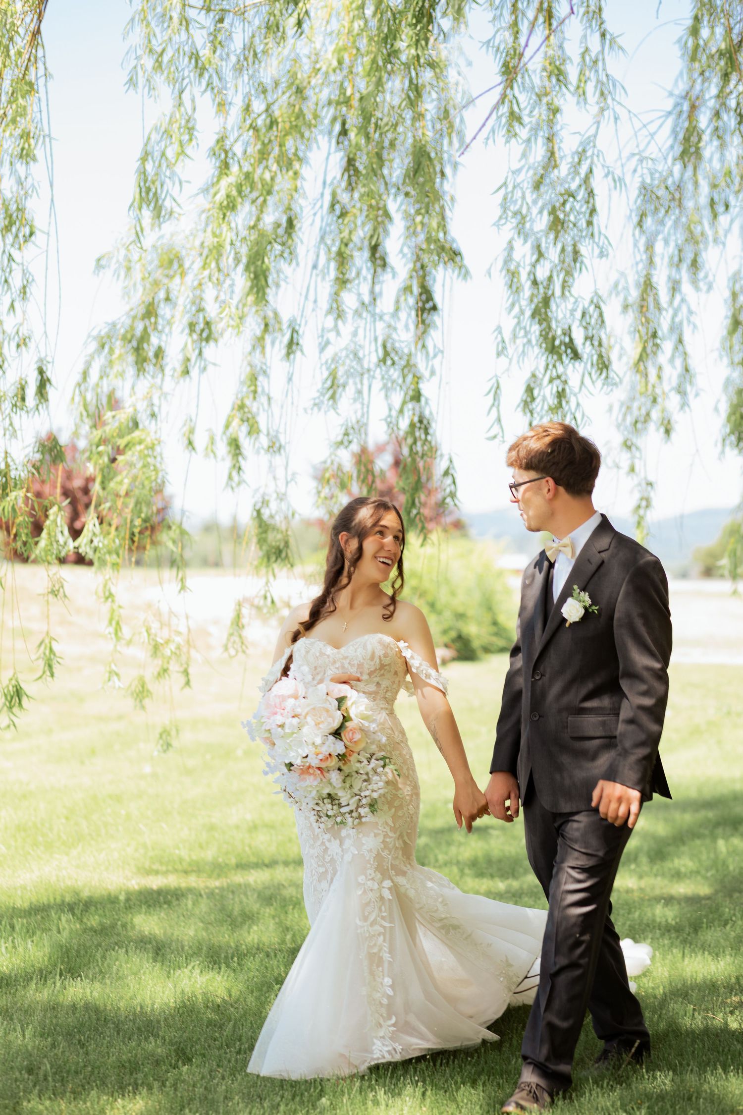 Bride and groom walk hand-in-hand under a weeping willow tree, bride holding a pastel bouquet in a lace mermaid gown.