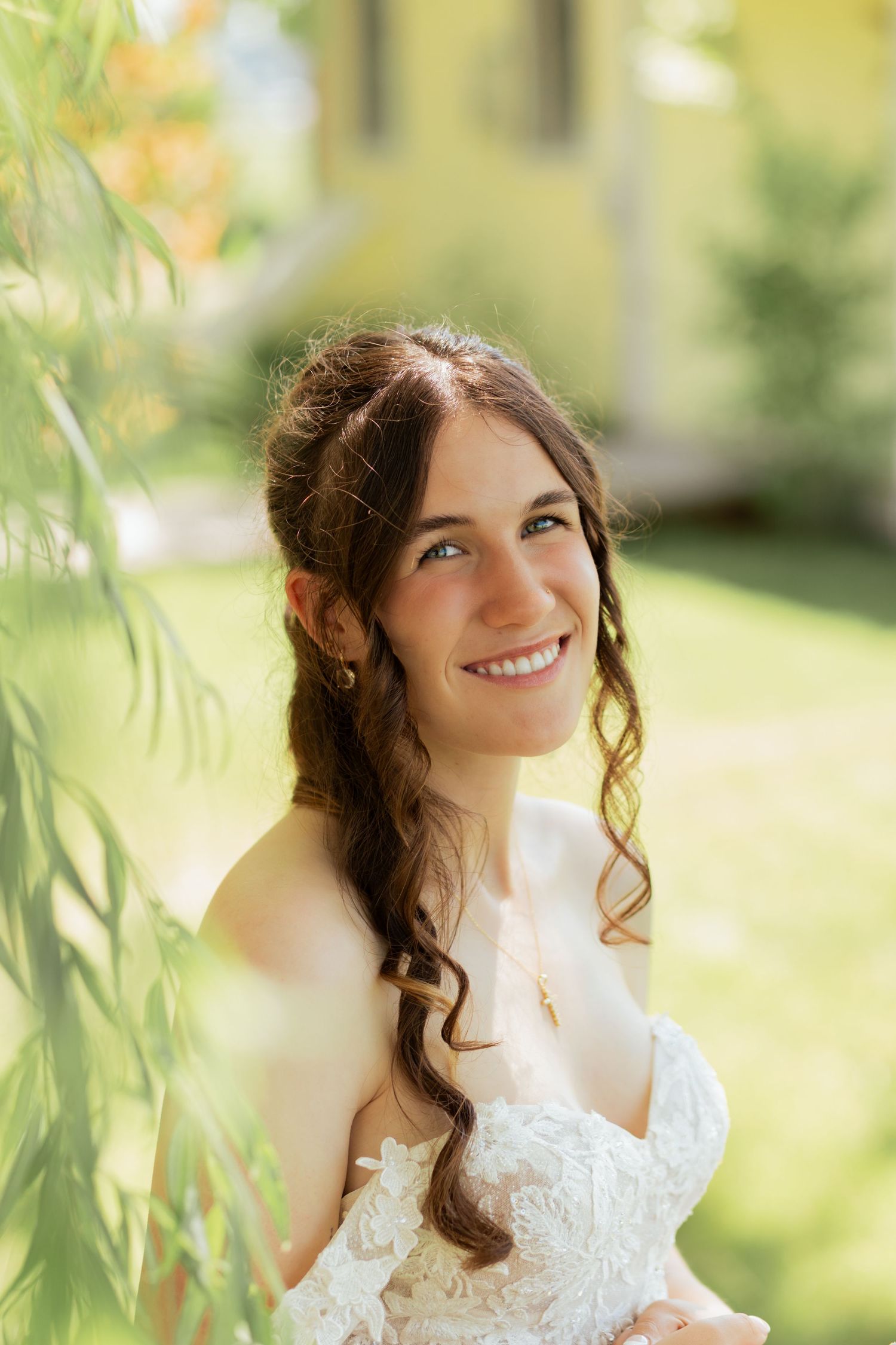 Smiling bride in white lace dress with curly hair poses outdoors in sunlit garden setting.