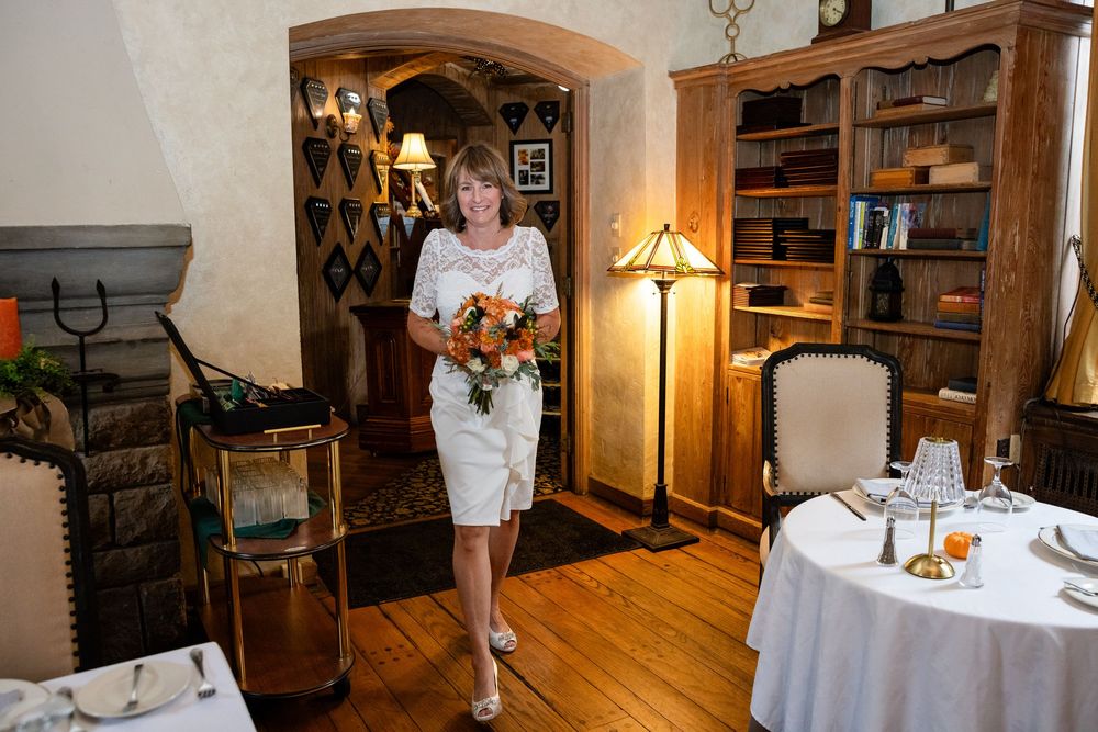Elegant bride in white dress carries a bouquet of orange flowers through a rustic restaurant wedding venue.