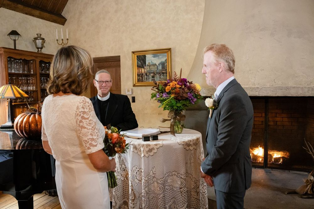 Bride and groom standing before officiant during intimate indoor wedding ceremony by fireplace.