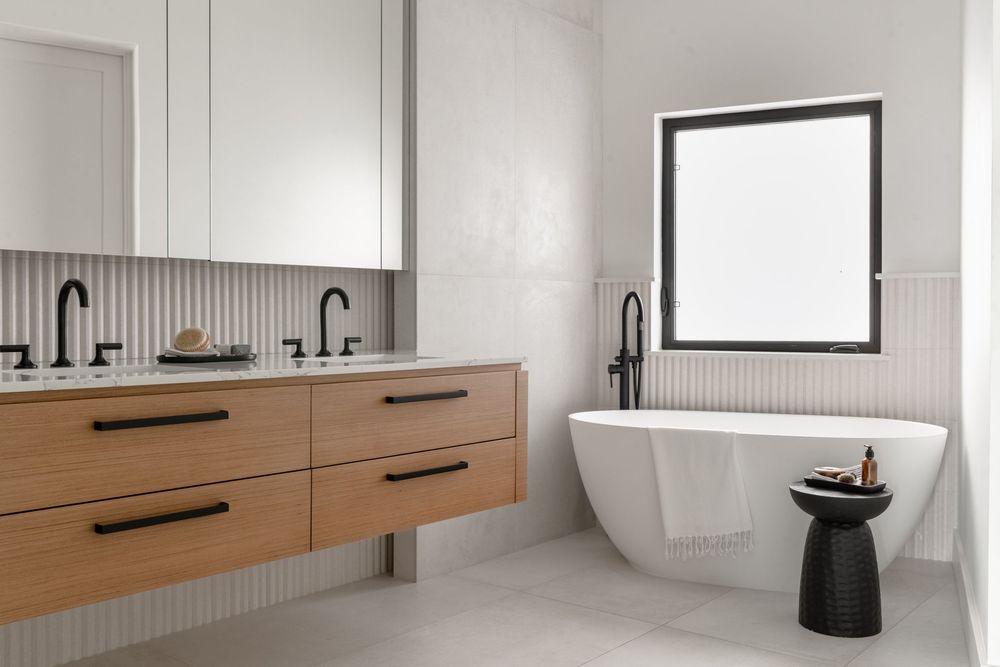 Modern white bathroom with floating wood vanity, black fixtures, freestanding soaking tub, and natural light from a square window.